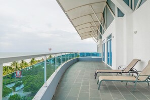 A balcony with two lounge chairs, a glass balustrade, and a view of the ocean and greenery.