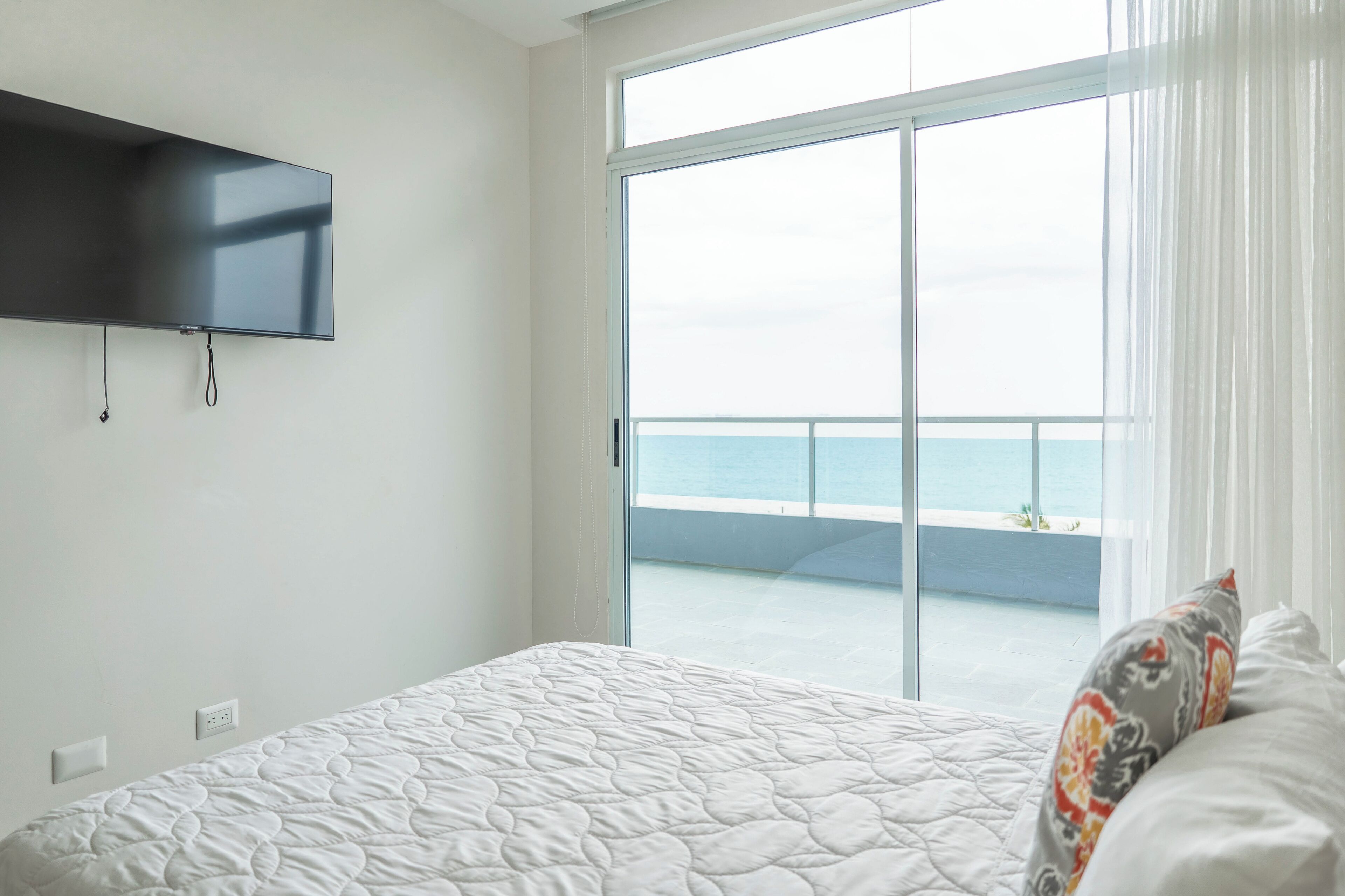 A bedroom with a bed, a television mounted on the wall, and a view of the sea through a sliding glass door.