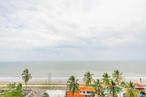A coastal view with a beach, palm trees, and buildings with orange roofs.