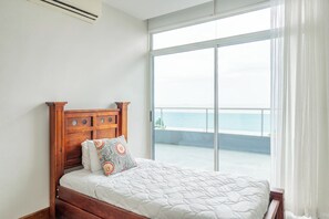 A bedroom with a wooden headboard, a bed with a white quilt, a patterned pillow, and a large sliding glass door leading to a balcony with a view of the sea.