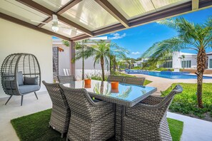 A modern outdoor seating area with wicker furniture and a glass table, overlooking a pool and palm trees. - Playa Avellanas Villa: Modern Luxury, Pool, Views (Playa Avellanas)