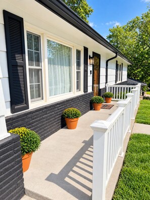 A two-story house with a front porch, black shutters, and potted plants.