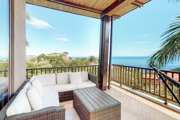 A balcony with a wicker sofa and a table, overlooking a coastal landscape with trees and a view of the sea.