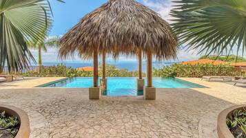 A pool area with a thatched structure, palm trees, and a view of the sea.
