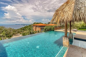 A clear blue infinity pool with a thatched umbrella, a view of the sea, and a building with a red roof. - Ocotal Hilltop Condo: Ocean Views, Pool Access (Playa Ocotal)