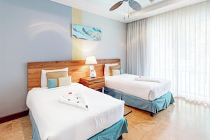 A hotel room with two beds, a wooden headboard, a nightstand with a lamp, and a large window with sheer curtains.