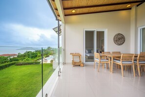 A balcony with a glass door leading to a bedroom, a dining table with chairs, and a view of a lush garden and distant buildings. - Ocean-View Tamarindo: Community Pool Access (Tamarindo)