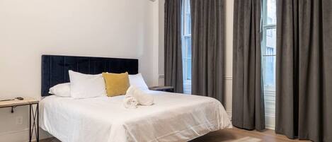 A hotel room with a bed, a bedside table, an air conditioning unit, and large windows with curtains.