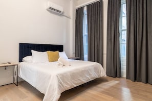 Et hotelværelse med seng, natbord, klimaanlæg og store vinduer med gardiner.