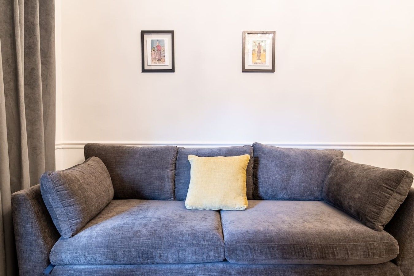 Ein graues Sofa mit einem gelben Kissen in einem Raum mit schlichten Wänden und zwei gerahmten Bildern.