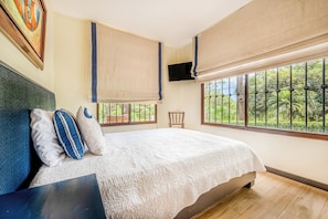 A bedroom with a large bed, a TV mounted on the wall, and a window with a view of greenery.