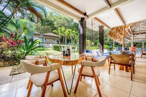 A covered outdoor seating area with a round wooden table and cushioned chairs, surrounded by lush tropical plants and a view of a house. - Private Pool, Luxe Patio: 4BR Carrillo Villa (Playas del Coco)