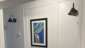 A hallway with white paneled walls, a framed artwork, a bench, and wall-mounted lighting fixtures.