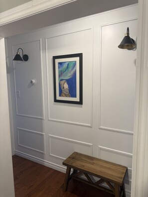 A hallway with white paneled walls, a framed artwork, a bench, and wall-mounted lighting fixtures.