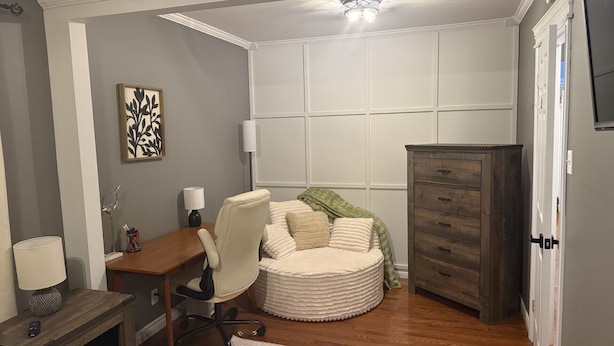 A compact room with a desk, chair, and a round ottoman with pillows.