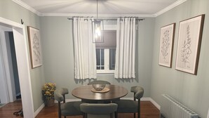 A dining area with a round table, four chairs, a window with curtains, and framed botanical prints on the wall. - Grand Falls-Windsor Gem | King Bed + Movie Room (Grand Falls-Windsor)