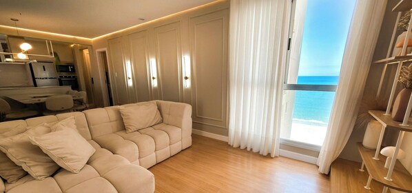 A modern living room with a beige sofa, a dining area, and a view of the ocean through large windows. - ASA 2 Suites Pe na Areia Vista mar BC (Balneário Camboriú)