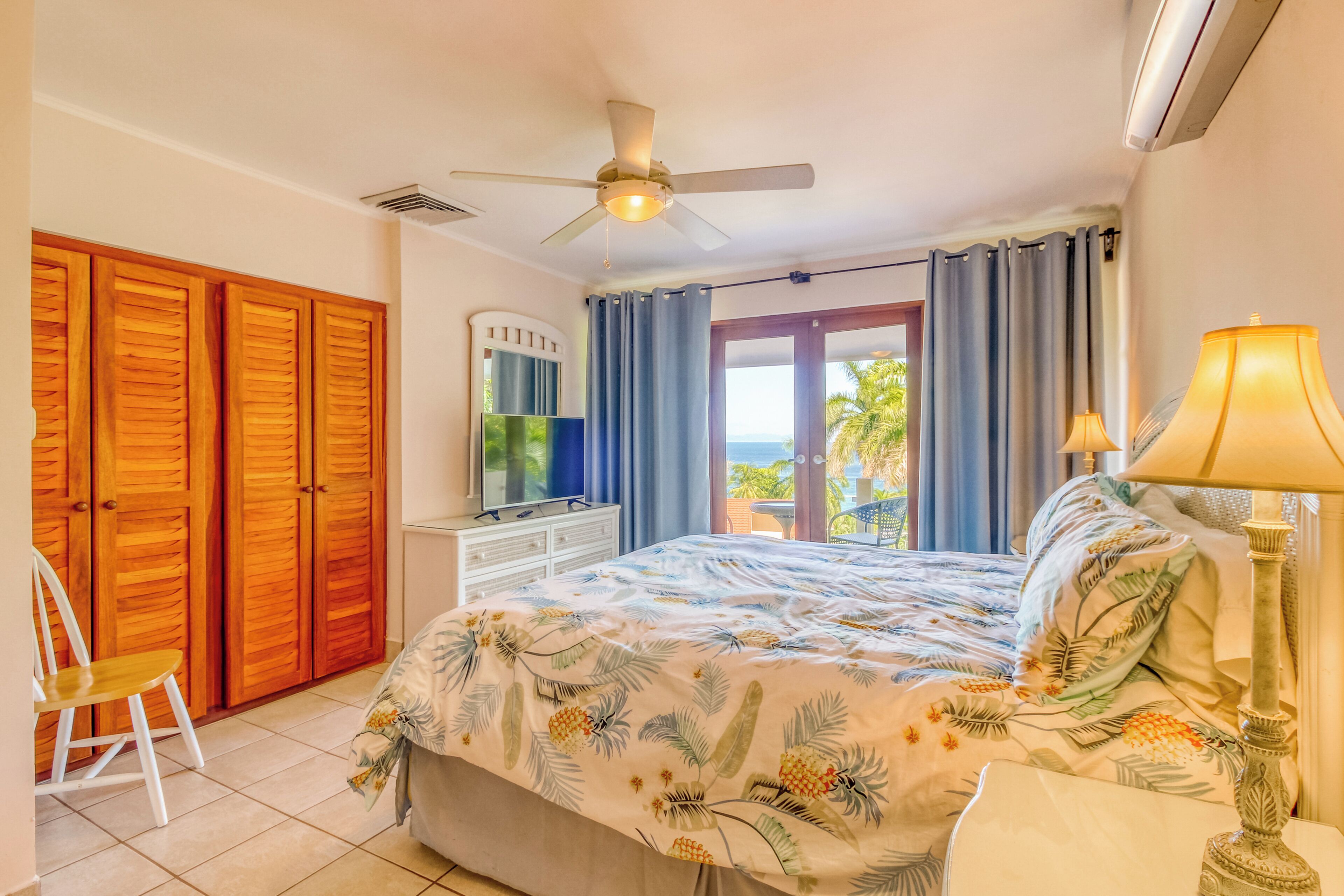 Un dormitorio con cama, televisor, ventilador de techo, ventana con cortinas y vista al exterior.