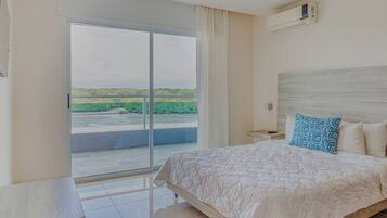 A hotel room with a bed, a desk, a chair, a window with a view of greenery, and an air conditioning unit.