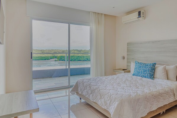 A hotel room with a bed, a desk, a chair, a window with a view of greenery, and an air conditioning unit.