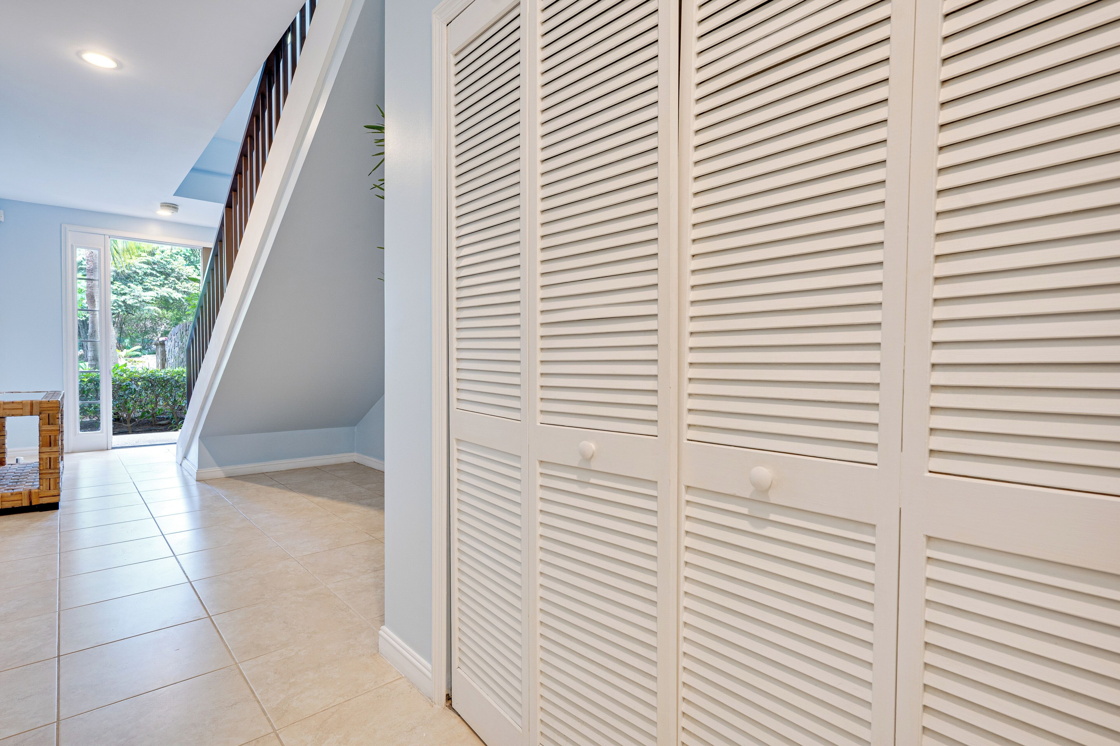 Un couloir avec un escalier menant à un étage supérieur, une armoire blanche dotée de portes à persiennes horizontales et un sol carrelé.
