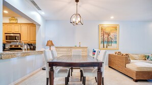 Uma área de refeições com mesa e cadeiras de madeira, uma cozinha com armários de madeira e um quadro com palmeiras na parede.