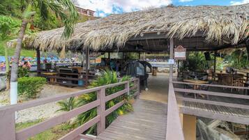 A covered outdoor seating area with wooden tables and chairs, a thatched roof, and a wooden walkway leading to it.