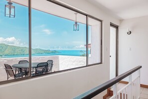 A balcony with a table and chairs, overlooking a coastal landscape with hills and the sea.