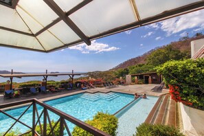 A swimming pool with clear blue water, surrounded by lounge chairs and umbrellas, with a view of the sea and hills in the background. - Playa del Coco 3BR: Ocean Views & Pools! (Playa Hermosa)
