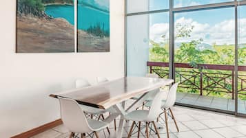 Un espace repas comprenant une table et des chaises, de grandes fenêtres donnant sur un paysage verdoyant et un bâtiment, ainsi que deux tableaux accrochés au mur.