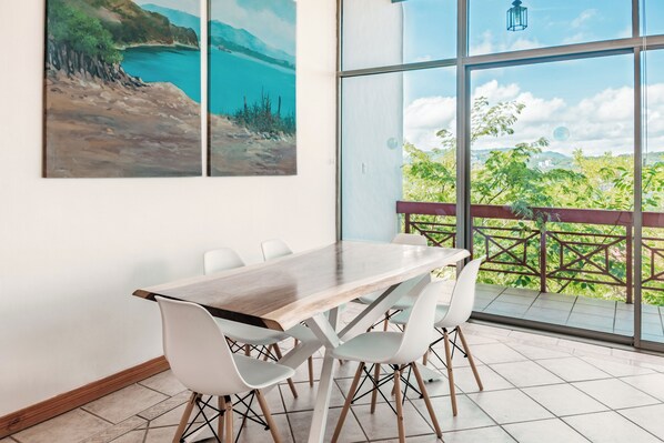 A dining area with a table and chairs, large windows with a view of greenery and a building, and two paintings on the wall.