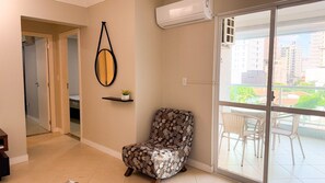 A living room with a chair, a wall-mounted mirror, a small shelf with a plant, and a sliding glass door leading to an outdoor area with wicker furniture. - ASA Sea View 2BR Apartment Pereque (Porto Belo)