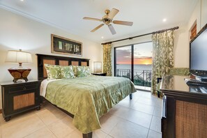 A hotel room with a bed, a nightstand, a lamp, a ceiling fan, a TV, and a view of the sunset through the window. - Best Flamingo Beach View! Pool, Balcony (Flamingo Beach)