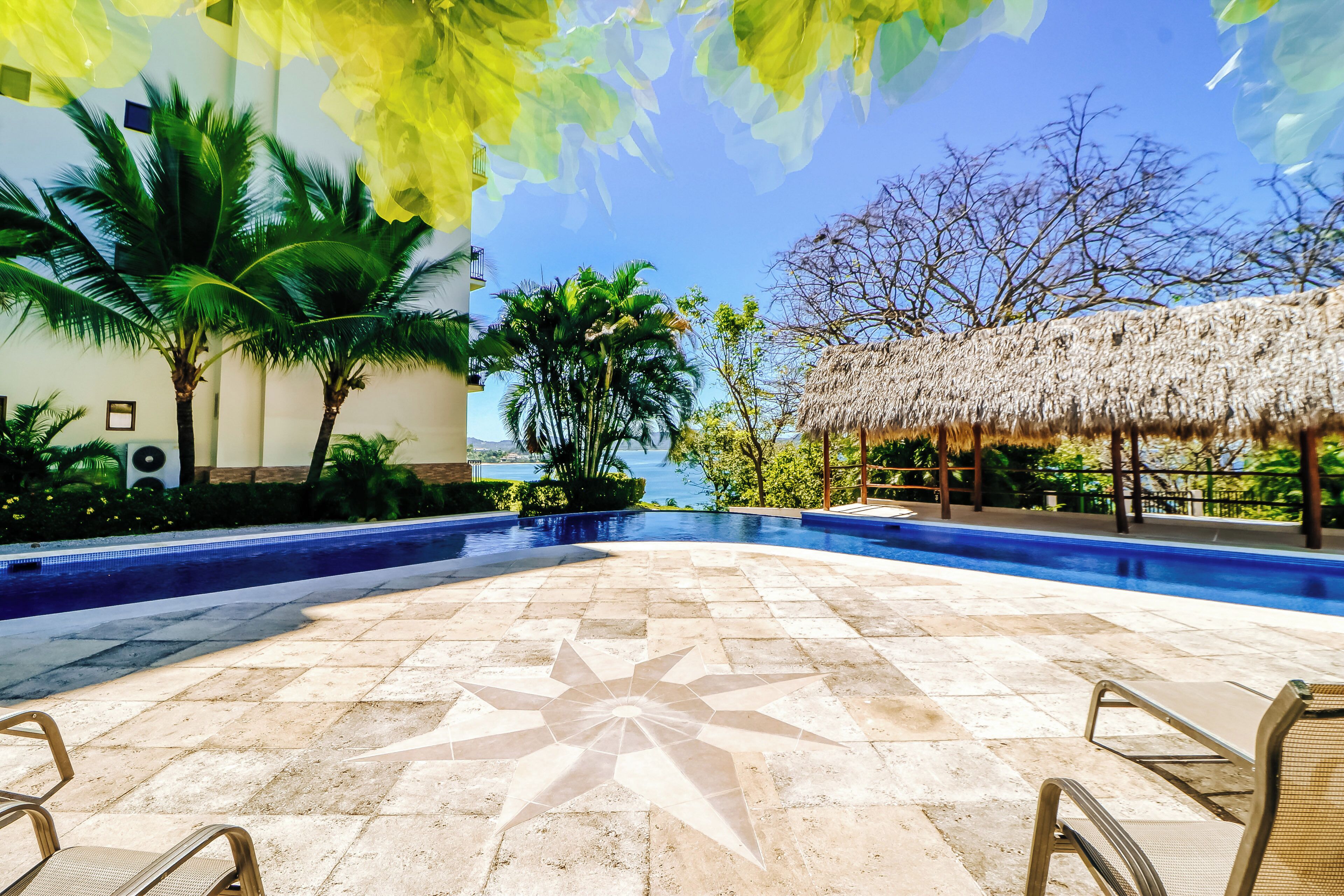 A swimming pool with a star-patterned tile design, surrounded by palm trees and a thatched-roof structure.