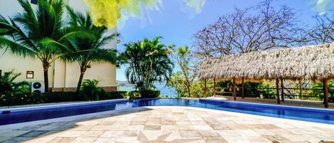 Una piscina con diseño de mosaicos formando un patrón de estrellas, rodeada de palmeras y una estructura con techo de paja.