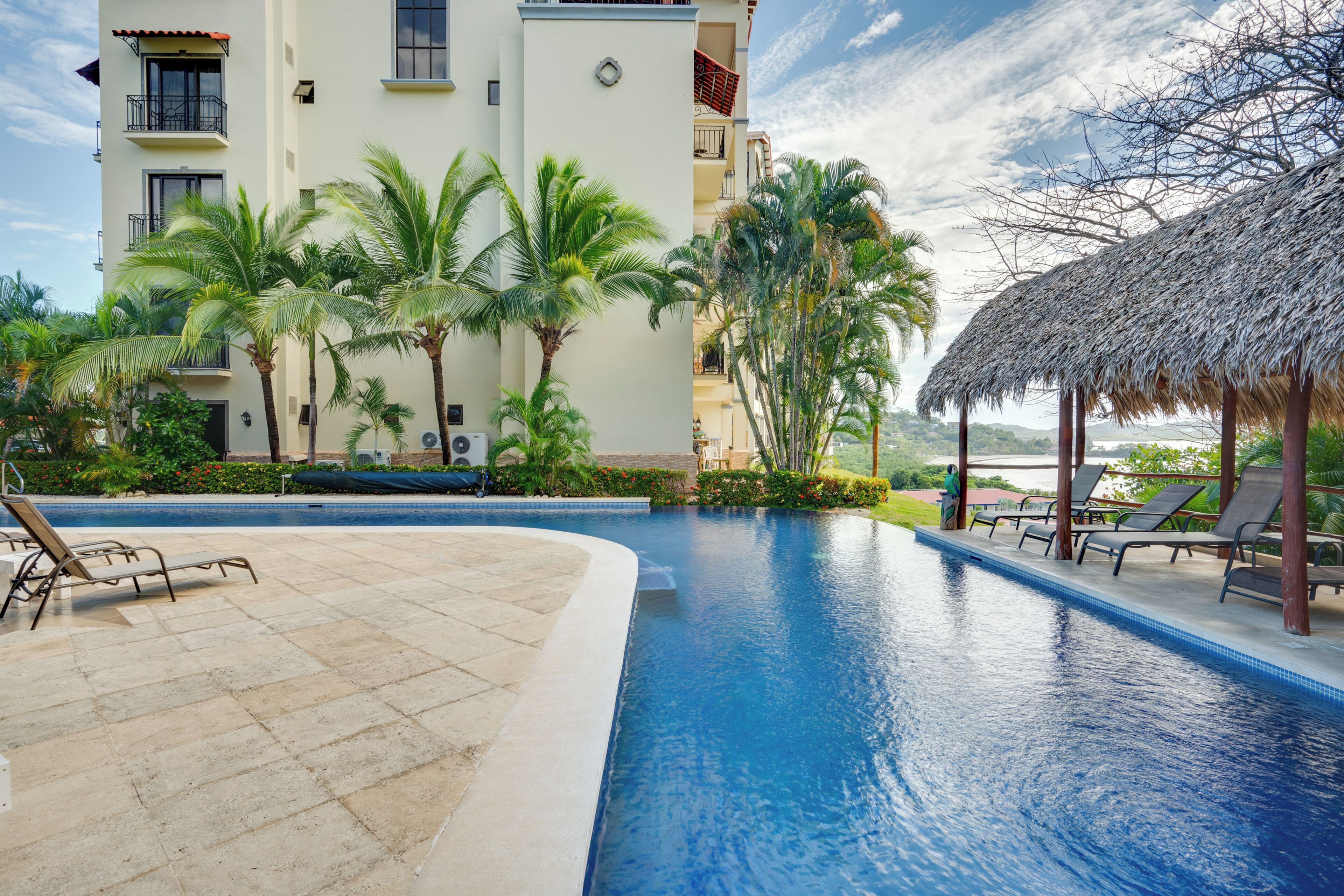 A swimming pool with lounge chairs, palm trees, and a building with balconies.