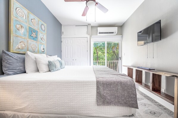 Une chambre avec un grand lit, un ventilateur de plafond, une télévision et une vue sur la verdure environnante.