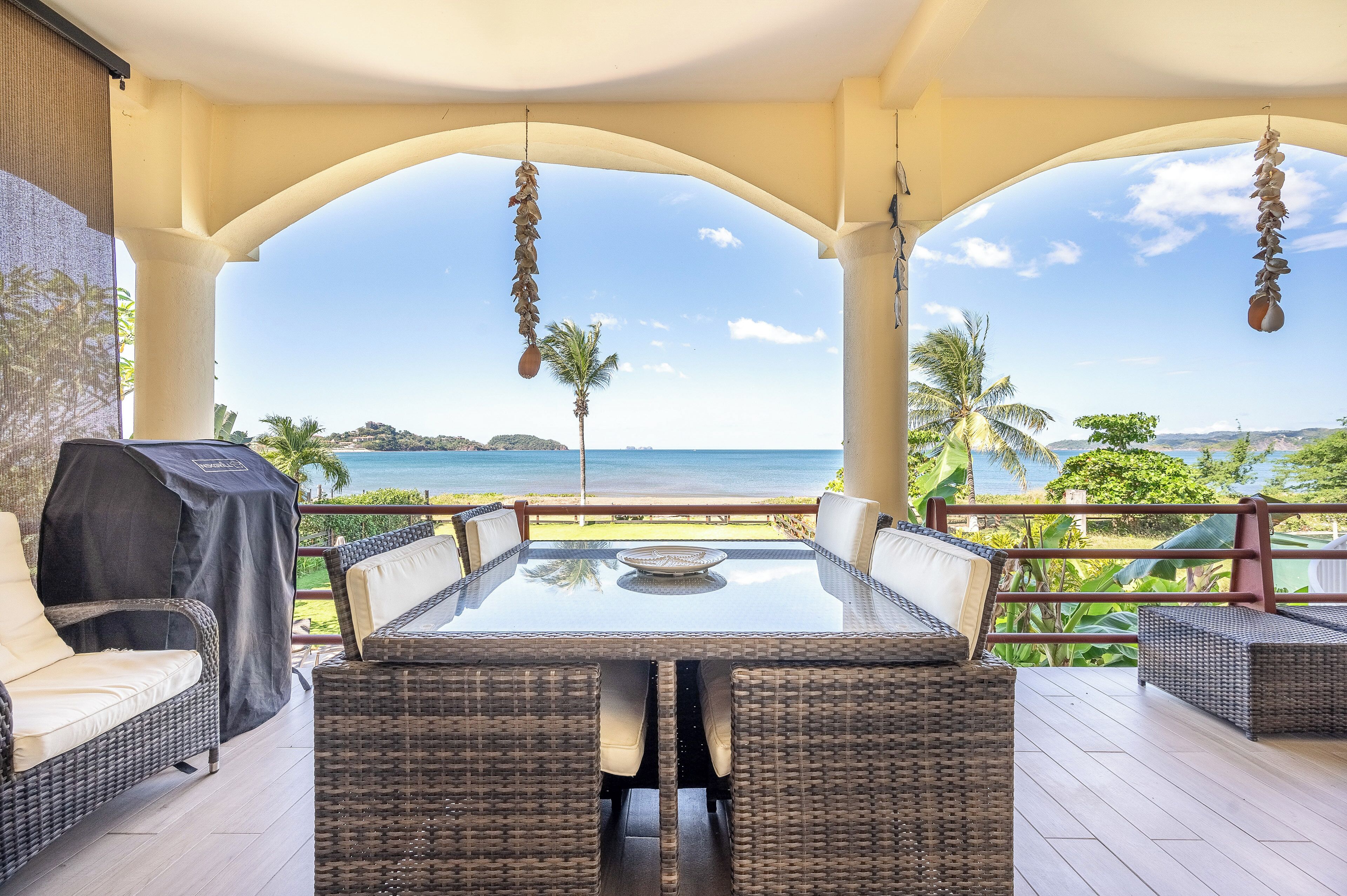 Une terrasse extérieure dotée d'une table à plateau de verre et de meubles en osier, offrant une vue sur une plage et l'océan.