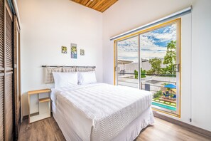 A bedroom with a bed, a nightstand, a window with a view of the outside, and a ceiling with wooden beams. - Walk to Del Coco Beach & Restaurants: 2 Condos! (Playas del Coco)