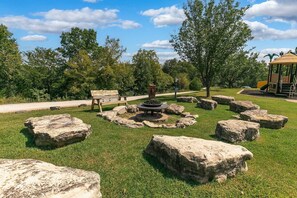 Ein Park mit einem runden Feuerkorb aus Stein, einer hölzernen Bank und einem Spielplatz im Hintergrund.