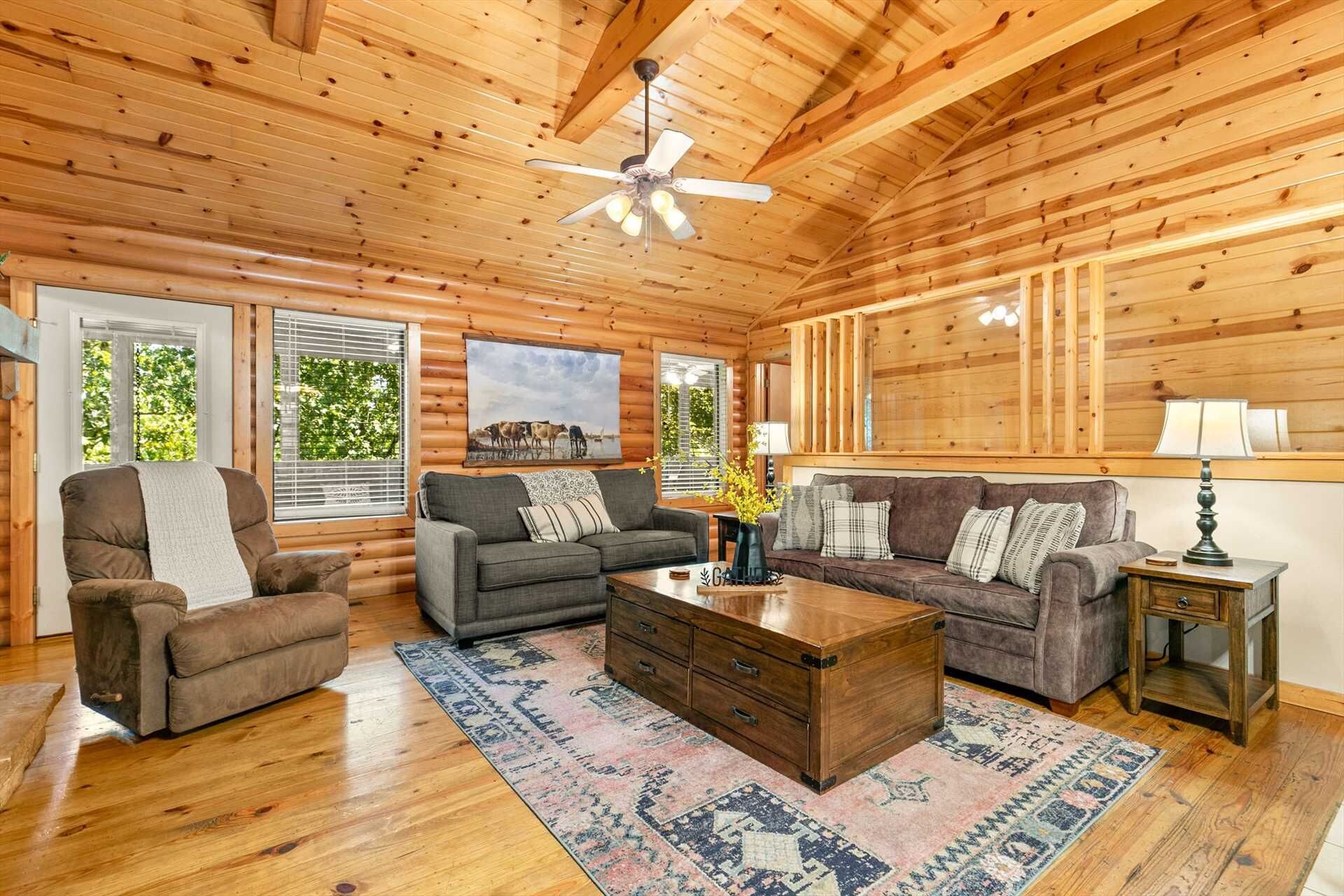 A log cabin interior with a wooden ceiling, two sofas, a coffee table, and a side table with a lamp.