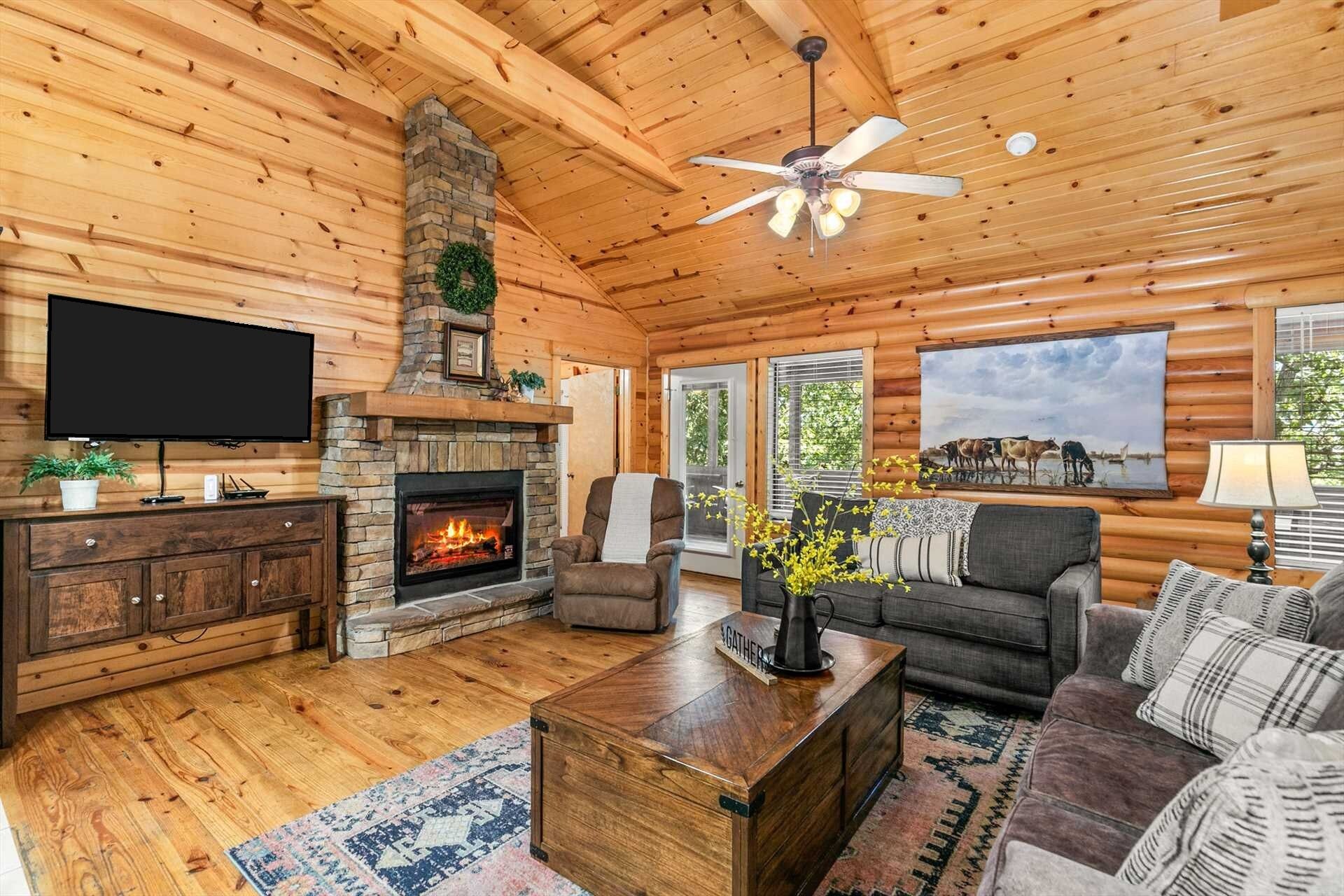 A log cabin interior with a fireplace, television, sofa, and a ceiling fan.