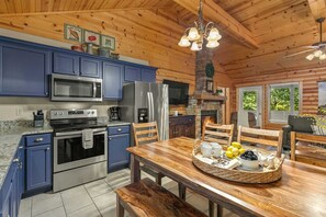 A kitchen with blue cabinets, stainless steel appliances, a wooden dining table with a fruit basket, and a stone fireplace. - Luxury in the Ozarks-Four  Master Bedroom Suites, Sleeps 14, Lots of Space for the Whole Family!-20 (Branson)