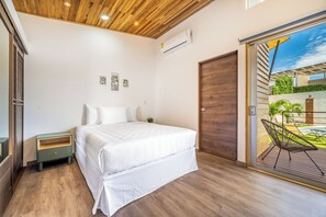A bedroom with a bed, a nightstand, a wooden ceiling, and a sliding glass door leading to an outdoor area with a pool and chairs. - Modern Studio w/ Pool Access Near Playas Del Coco! (Playas del Coco)