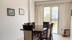 Un comedor con mesa de vidrio y cuatro sillas, una puerta corrediza de vidrio a un balcón y dos cuadros en la pared.
