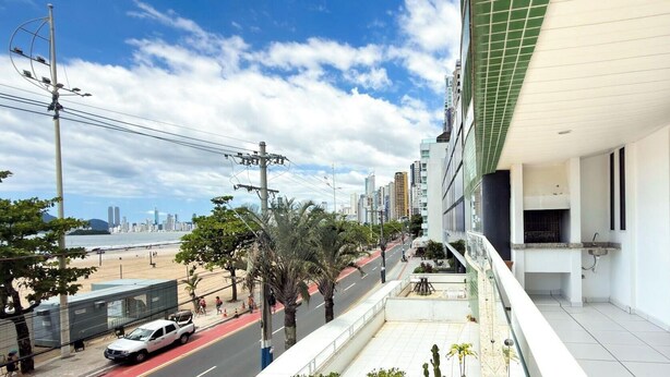 Un balcón con vista a la playa y al horizonte de la ciudad, un coche blanco en la calle, y un edificio con tejado de tejas verdes.