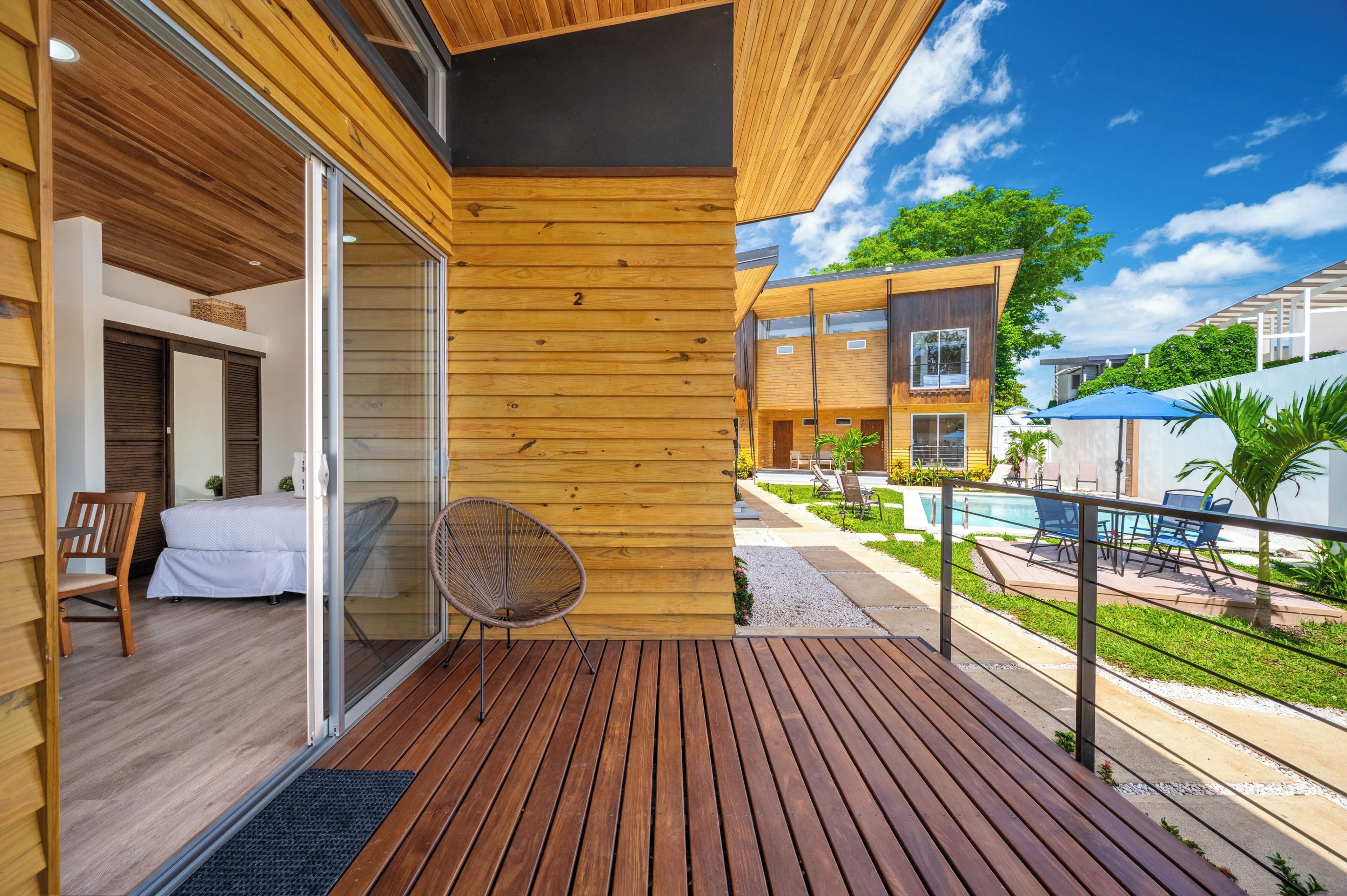 A wooden deck with a glass door leading to a bedroom, a lounge chair, and a pool area with umbrellas.