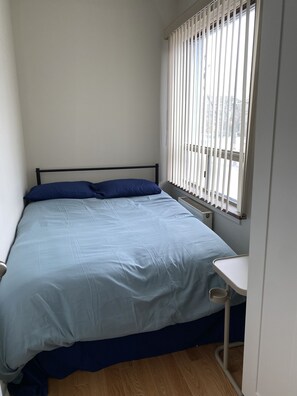 Een slaapkamer met een bed, een raam met zonwering, een nachtkastje en een radiator.
