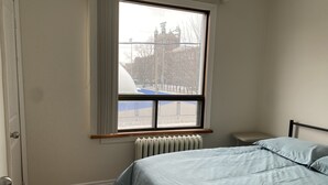 Quarto com uma cama de solteiro, radiador, janela com vista para um edifício e porta.