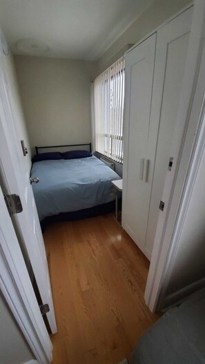 Een kleine kamer met een enkel bed, houten vloer, raam met zonwering en een kledingkast.